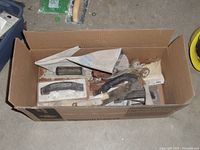 Photo of a cardboard box filled with various masonry and concrete hand tools including a pointed trowel, a wide trowel, and a brush, all showing signs of use.