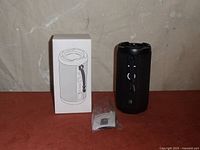 Cordelia IT151 Bluetooth speaker beside original box and small plastic packet containing USB cable or instructions, placed on a surface against a plain wall background.