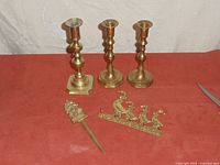 Full lot showing three candlesticks, brass letter opener, and brass key hook on red surface with some scissors visible at edge.