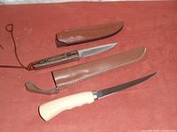 Two bushcraft and fishing knives placed on a reddish surface, showing blade, handle, and leather sheath of each knife.
