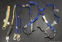 Top-down photo showing the blue and grey Norguard universal fit safety harness alongside three lanyards: two blue elastic lanyards with large metal hooks and one yellow fabric lanyard with a smaller metal hook.