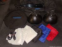 Photo showing two metal reflector photo lights, cords, folded white fabric backgrounds, four colored fabric sheets (white, black, blue, red), and a black round storage bag with Inspiron logo.
