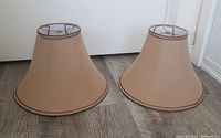 Two matching taupe/sand brown bell-shaped lamp shades sitting on wood floor showing front and side profiles.