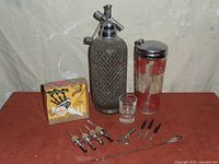 Full lot display showing vintage barware including soda siphon dispenser, glass container with silver lid and floral pattern, shot glass, boxed musical bottle stopper, and various bar accessories.