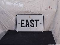 Front view of rectangular metal road sign in white with bold black letters spelling 'EAST'. Sign has rounded corners and visible mounting holes.