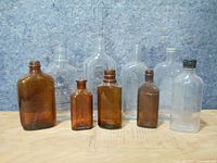 Photo showing eight vintage glass liquor bottles arranged in two rows; four amber bottles in front and four clear bottles behind on a wooden surface with a blue textured background.