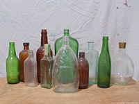 Group of 6 vintage glass bottles of different colors and shapes, displayed on floor against wall.