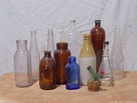 7 vintage glass bottles arranged on a textured white backdrop; includes clear, amber, cobalt blue, and two-tone ceramic bottles along with a miniature barrel decorative item.