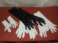 All four pairs of gloves laid out on rust-colored surface, showing black, two pairs of long white gloves, and one smaller white pair.