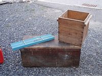 Photo showing the two wooden storage items: the dark wood toolbox lying on the ground with its hinged lid closed, and the lighter wood crate placed on top of the toolbox.