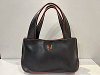 Front view of black Armani Jeans handbag with embroidered orange AJ initials and orange edge piping.