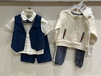 Photo showing two baby boy clothing sets hanging on a wall. Left is a Jean Bourget 4-piece set with navy vest, white shirt, shorts, and bow tie. Right is Baby Mode 2-piece sweat suit with cream textured sweatshirt and gray sweatpants, both with price tags.