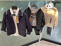 Photo of three baby boys clothing sets hanging on a rack: navy blazer set, gray positive vibes set, cream knit set with pants.