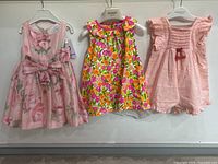 Three baby girl clothing items hanging side by side: pink floral dress, magenta and orange floral dress, pastel pink romper.