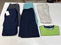 Six pairs of boys pants folded on a white surface along with a folded lime green shirt with a blue collar. Pants colors include navy, gray, light blue, and off-white.