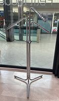 Silver metal clothing display rack with three arms, each adjustable in height and angle, on a tri-pod base standing on tiled floor near glass window.