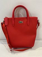 Front view of red Lacoste purse showing green crocodile logo, dual handles, strap, and zipper pull.