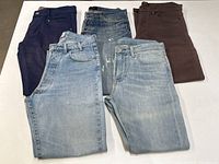 Five pairs of men’s jeans and pants laid flat showing varied colors including light blue denim, dark blue/purple, medium wash blue, and brown.