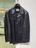 Full front view of the black leather jacket hanging on a Versace hanger showing the classic design and button closure.