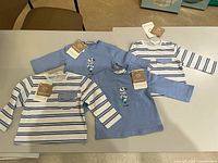 Photo showing all four newborn cotton tops laid out on a table: two solid blue with graphic prints and two white with blue stripes and chest pocket, all tagged.
