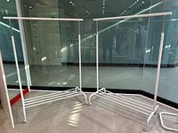 Two white adjustable rolling clothing racks side by side in a corner of a room with reflective glass walls.