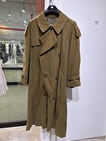 Full view of the olive green trench coat hanging showing double-breasted button closure, length and collar design.