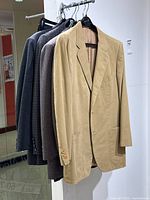 Five vintage blazers hanging on black hangers on a metal rack, showing various colors and fabrics including beige, brown, and gray.