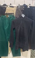 All 6 clothing pieces hanging on hangers: black DDP capri pants with flap pockets, beige CHEVIGNON pants, dark grey VERSACE pants, black MISS SIXTY pants, green CAMPUS pants, and black TALULA hoodie at front.