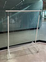 White metal rolling clothing rack with adjustable height, shown from an angle in front of a glass wall