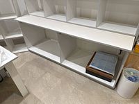 Front view of white melamine cabinet/workbench showing two large open cubbies with vertical divider and flat top surface with visible wear and holes.