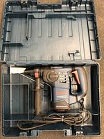 Top view of Bosch RH328VC rotary hammer in black and blue, with red triggers, inside molded hard plastic case. Auxiliary handle attached to front side of tool. Coiled power cord included in case.