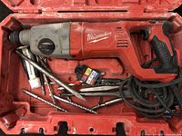Rotary hammer drill with attached power cord inside the open red plastic case, several drill bits and a tube of lubricant also visible inside the case.