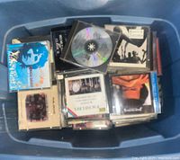 Blue plastic tub filled with assorted music CDs, displaying a variety of cases and some visible artist names.