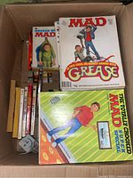 Box filled with various MAD magazines and books, some visible titles include 'Hooked on MAD' and MAD magazine featuring a Grease cover.