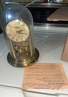 Photo of front side showing clock face with roman numerals and rotating pendulum balls, placed on a surface with instruction sheet visible