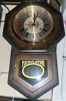 Front view of antique regulator wall clock showing octagonal face, black and red numbers, brass bezel, and lower door with "REGULATOR" decal.