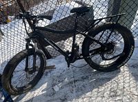 Full side profile of black Daymak Wildgoose electric fat-tire bike leaning against chain-link fence on snow