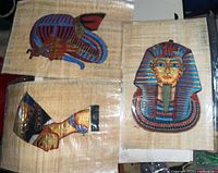 Two Egyptian papyrus prints, one with a colored sarcophagus mask and the other showing two Egyptian figure profiles in vibrant traditional colors, all protected in plastic.