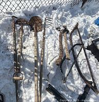 Photo showing seven garden tools including axes, spade, pitchfork, saws and hook placed on snow with a fence in the background.