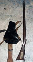 Photo showing the wooden rifle and black leather gun holster with brown strap side by side on a floor.