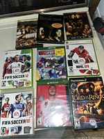 Photo showing three DVD movies: The DaVinci Code, Creation of the DaVinci Code, and The Mummy Returns, plus several video games including FIFA Soccer 12 Xbox 360, Transformers Devastation Xbox One, NHL 09 Xbox 360, FIFA Soccer 10 PS3, Xbox One FIFA 20 unopened, and The Lord of the Rings PS2.