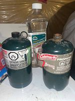 Photo showing two small green propane tanks and bottle of Coleman lamp oil on a white surface with plastic-covered wall background.