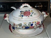 Side view of covered porcelain soup/stew pot with floral design and decorative handles, placed on matching underplate.