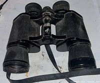 Single older style pair of black binoculars with leather strap, visible wear on the surface
