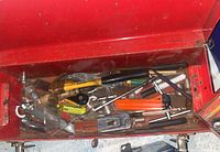 Top view inside of red metal toolbox showing assortment of hand tools including claw hammer, screwdrivers, chisels, socket wrench bits and extensions scattered inside.