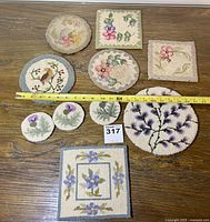 Overview of nine petit point trivets and coasters arranged on wood surface, showing size scale with tape measure and lot number card.