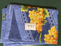 Folded blue cotton tablecloth with yellow grapes and green leaves with a paper label showing lot number 822 on top.