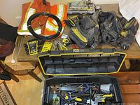 Photo showing Dewalt tool rig with multiple tool belts and pouches, orange high-visibility coveralls, black pressure line hose coiled, Stanley wrench sets, screwdriver bits and hand tools on table.