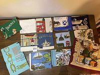 Full lot of 13 printed Irish linen tea towels neatly folded with various prints visible including green towel with Sailing Terms and blue with silver teapot.