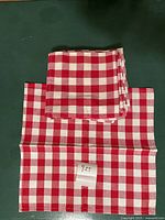 Single folded red and white gingham cloth napkin with paper price tag showing $25, on green surface.